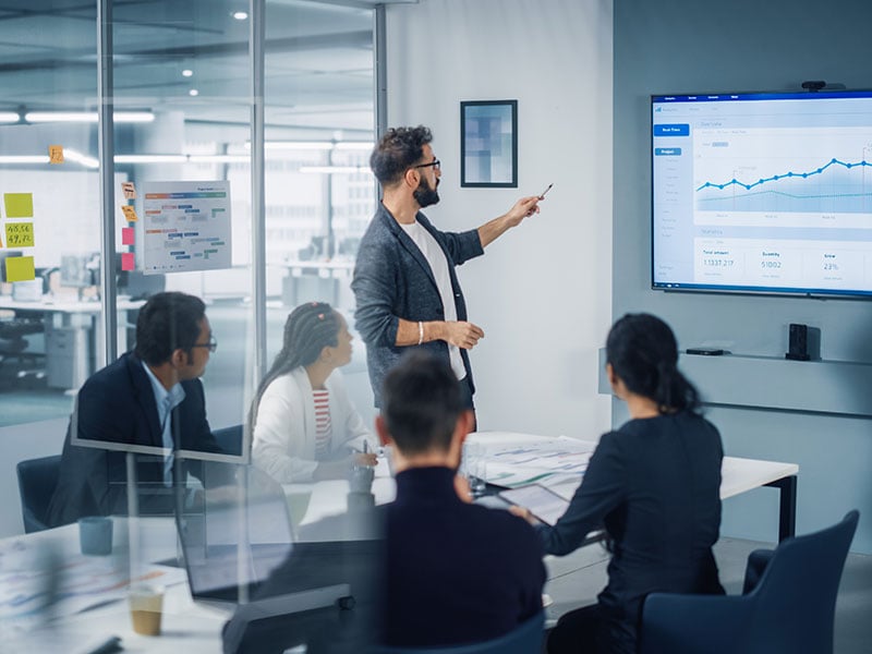 A businessman points at a presentation slide as four colleagues look on